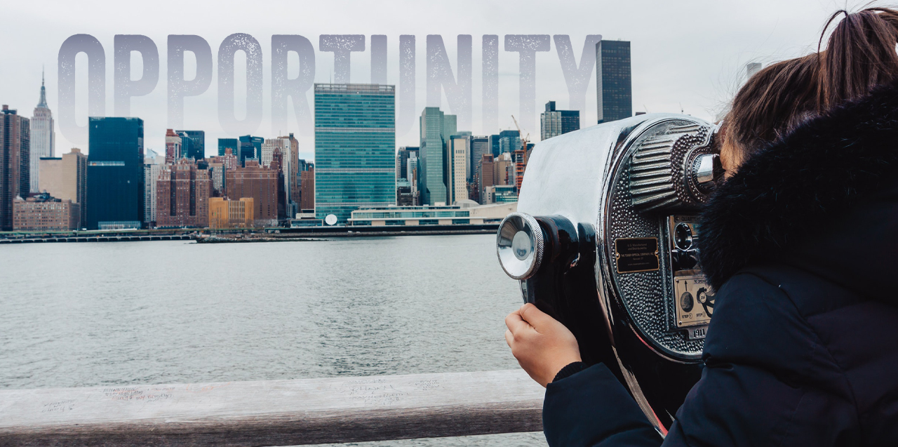 A person in a black fluffy coat with brown hair in a ponytail is looking through coin-operated binoculars across a body of water at a city skyline. Over the city the word "Opportunity" is superimposed on the sky.