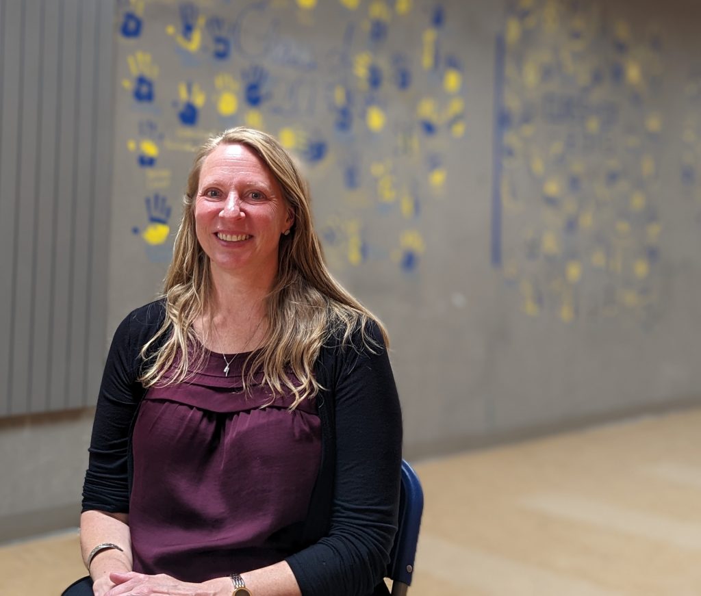 Photo of Michelle Eagleson sitting and smiling in front of a hallway wall that has the blue and yellow painted handprints of former graduated students.